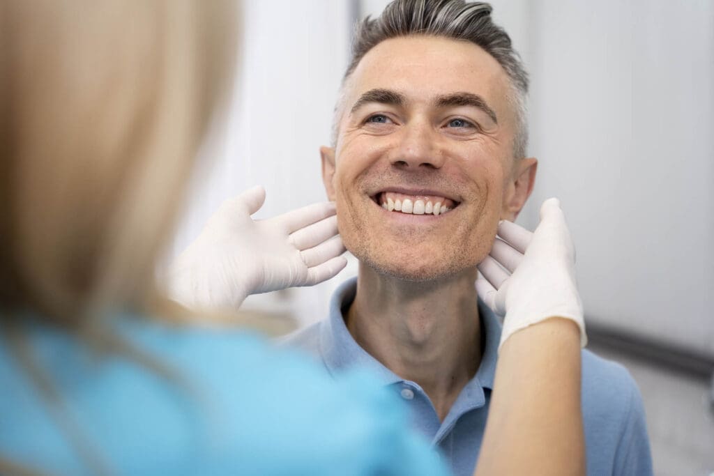Okemos Dentist A smiling man with short gray hair is examined by an Okemos dentist wearing white gloves, who is gently touching his chin and jaw. The man appears happy and relaxed during the checkup.