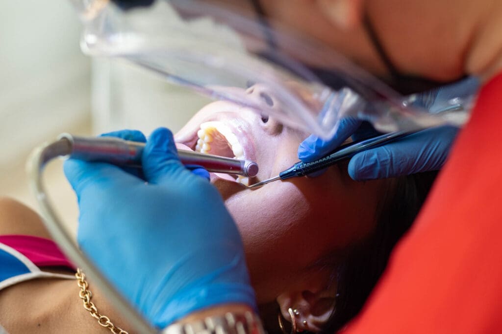 Okemos Dentist A close-up of a person receiving dental treatment, showing gloved hands using dental instruments inside the patient’s open mouth. The patient is lying back and wearing a protective bib.