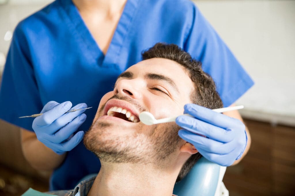 Okemos Dentist A person sits in a dental chair, smiling with their mouth open, while an Okemos dentist in blue scrubs and gloves examines their teeth using dental tools.