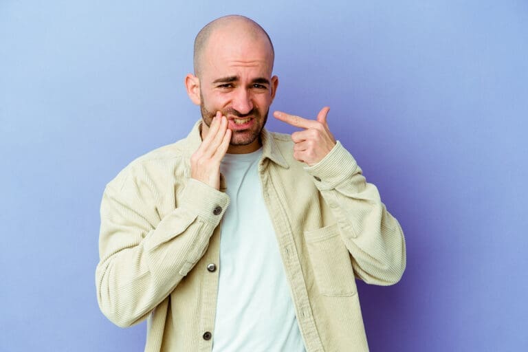 Okemos Dentist A man with a bald head and beard touches his cheek and points to his mouth in pain, standing against a purple background—possibly in need of care from an Okemos dentist.