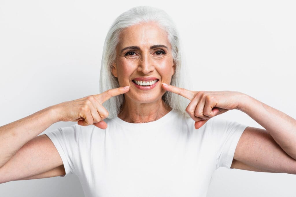 Okemos Dentist Smiling older woman with long gray hair in a white shirt points to her teeth with both index fingers, standing in front of a plain white background, showcasing her healthy smile after visiting an Okemos dental clinic.