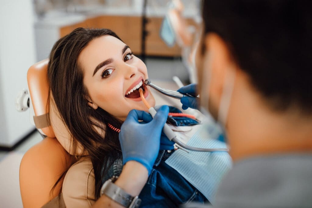 Okemos Dentist A woman reclines in a dental chair, smiling while an Okemos dentist wearing gloves examines her teeth with instruments. The dentist's face is out of focus in the foreground.