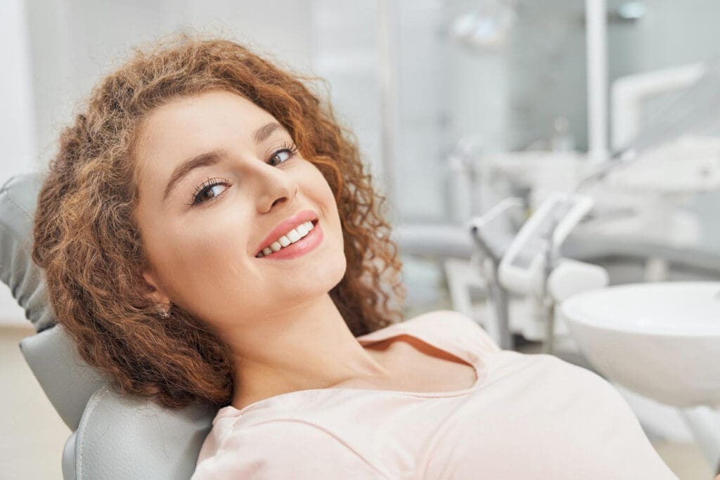 Okemos Dentist A young woman with curly hair smiles while sitting in a dental chair at an Okemos dentist's office, wearing a light-colored top. Dental equipment and a sink are visible in the background.