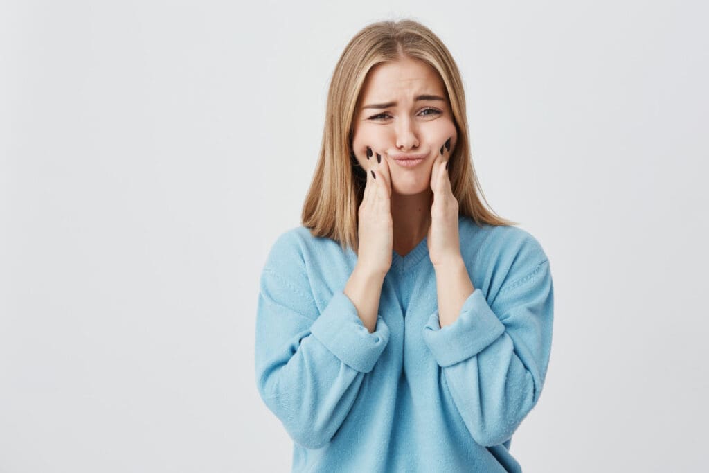 Okemos Dentist Young woman with long blonde hair in a light blue sweater presses her cheeks with both hands and makes a pouty, displeased expression, possibly needing an Okemos dentist, against a plain white background.