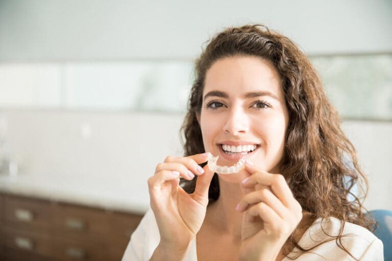 Okemos Dentist A smiling woman with curly hair holds a clear dental aligner near her mouth, showing her teeth in a bright, modern Okemos dental clinic.