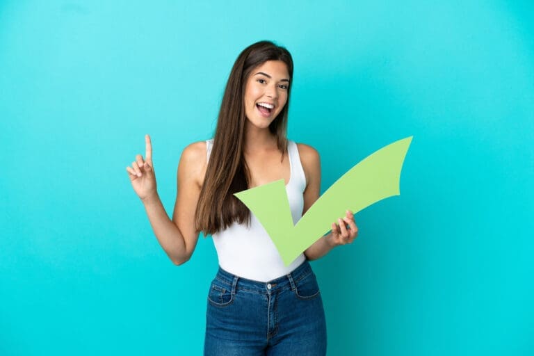 Okemos Dentist A smiling young woman with long brown hair stands against a blue background, holding a large green check mark cutout and pointing upwards. She appears positive and enthusiastic, embodying the friendly atmosphere of an Okemos dental clinic.