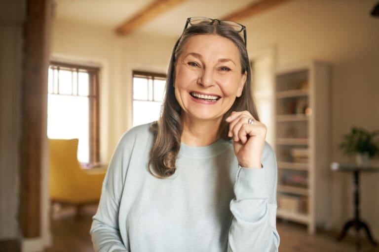 Okemos Dentist A smiling older woman with long gray hair wears glasses on her head and a light blue sweater, standing in a bright, cozy room at an Okemos dental clinic with large windows and shelves in the background.