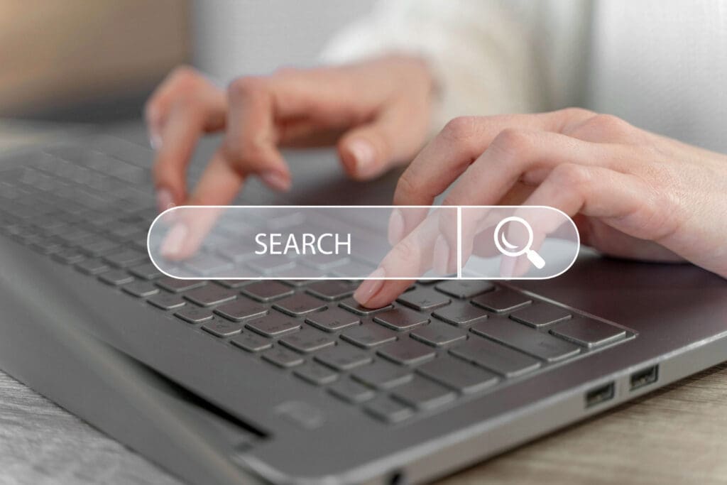 Okemos Dentist Close-up of a person’s hands typing on a laptop keyboard, with a transparent search bar graphic overlay displaying the word SEARCH and a magnifying glass icon.